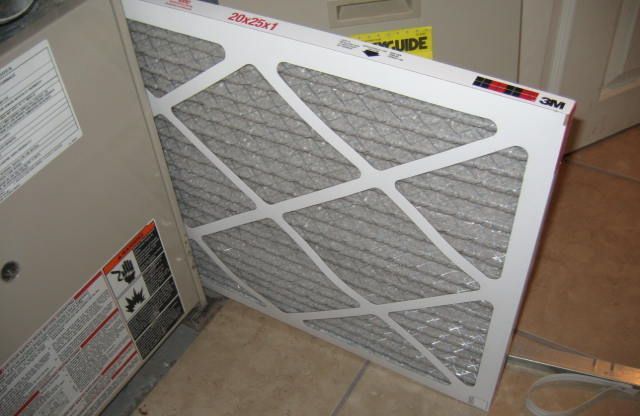 A rectangular air filter is held up next to an HVAC unit. Filter is light gray with white frame.
