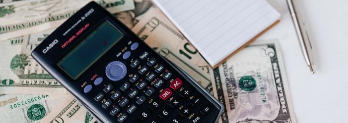 A calculator on the top of a pile of money and a notebook emphasizing the idea of crafting a perfect budget.