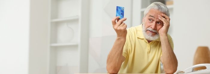 An elderly man is sitting at a table holding a credit card wondering What is Credit Card Fraud.
