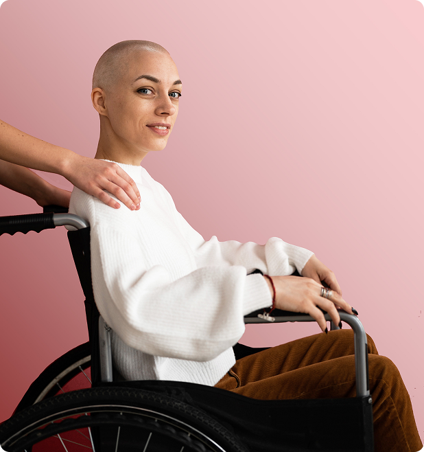 Woman in wheelchair with shaved head, smiling, with hand on shoulder, pink background.