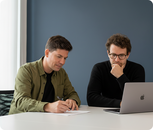 Two men at table, one writing, the other looking at laptop screen, in a blue-walled office.