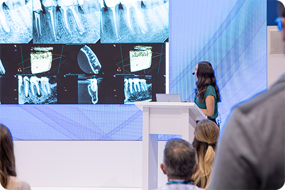 Woman giving presentation on dental x-rays. Audience watches; large screen displays various x-ray images.