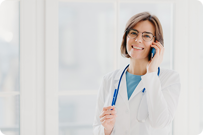 Doctor wearing a white coat and glasses smiles while talking on a phone, stethoscope around her neck.
