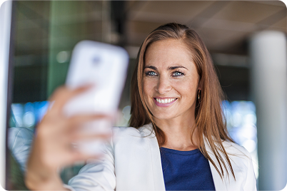 Woman taking a selfie with a smartphone, smiling. Wearing a white jacket and blue top, in a building.
