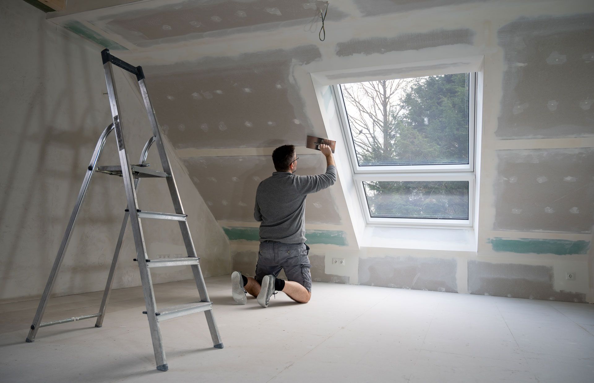 A man is kneeling down in front of a window in a room with a ladder.