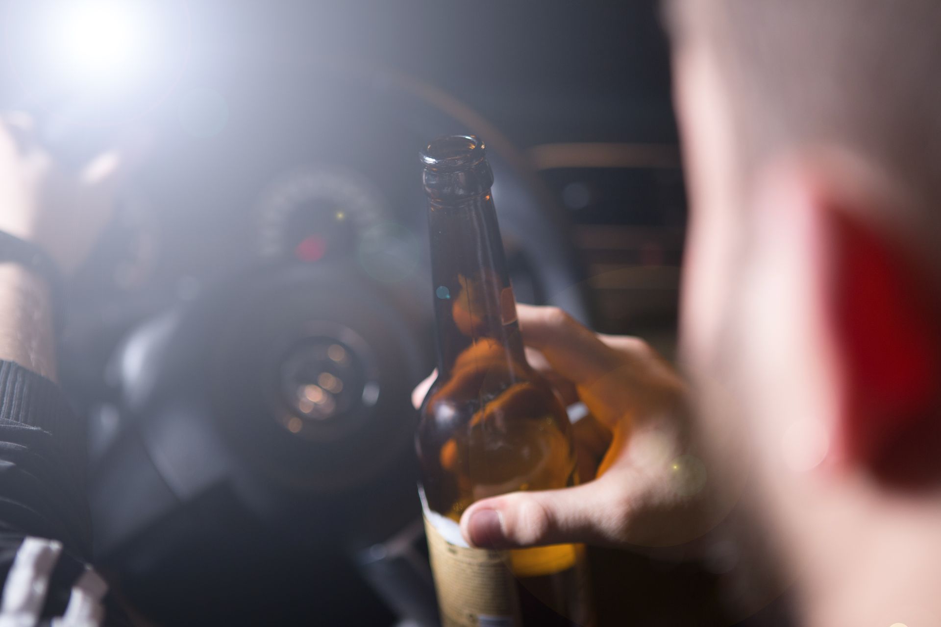Person holding beer bottle in car, approaching steering wheel. Bright light from dash.