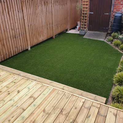 A square lawn next to wooden decking and surrounded by plants and a wooden fence
