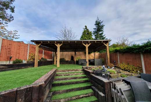 A wooden structure set on a lawn at the top of some grassy steps in a garden with wooden fencing around it