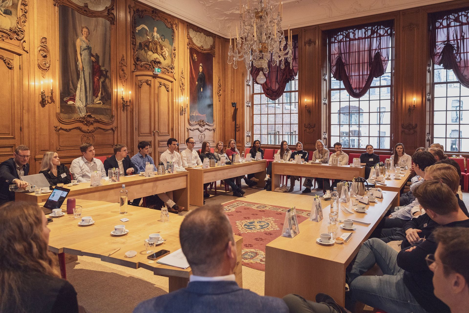 Een groep mensen zit rond een grote tafel in een sierlijke ruimte en discussieert met behulp van documenten, water en een aantal laptops.