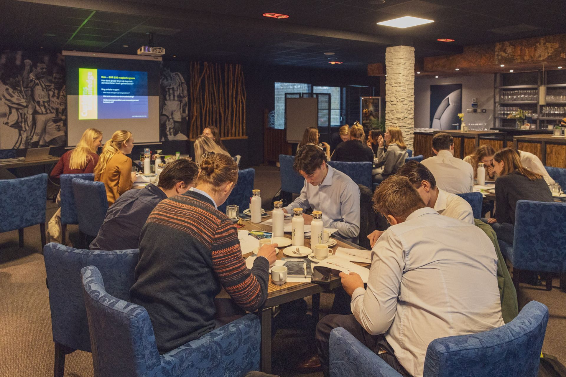 Mensen in een vergaderzaal; velen zitten aan tafels te werken. Op de achtergrond is een presentatiescherm te zien.