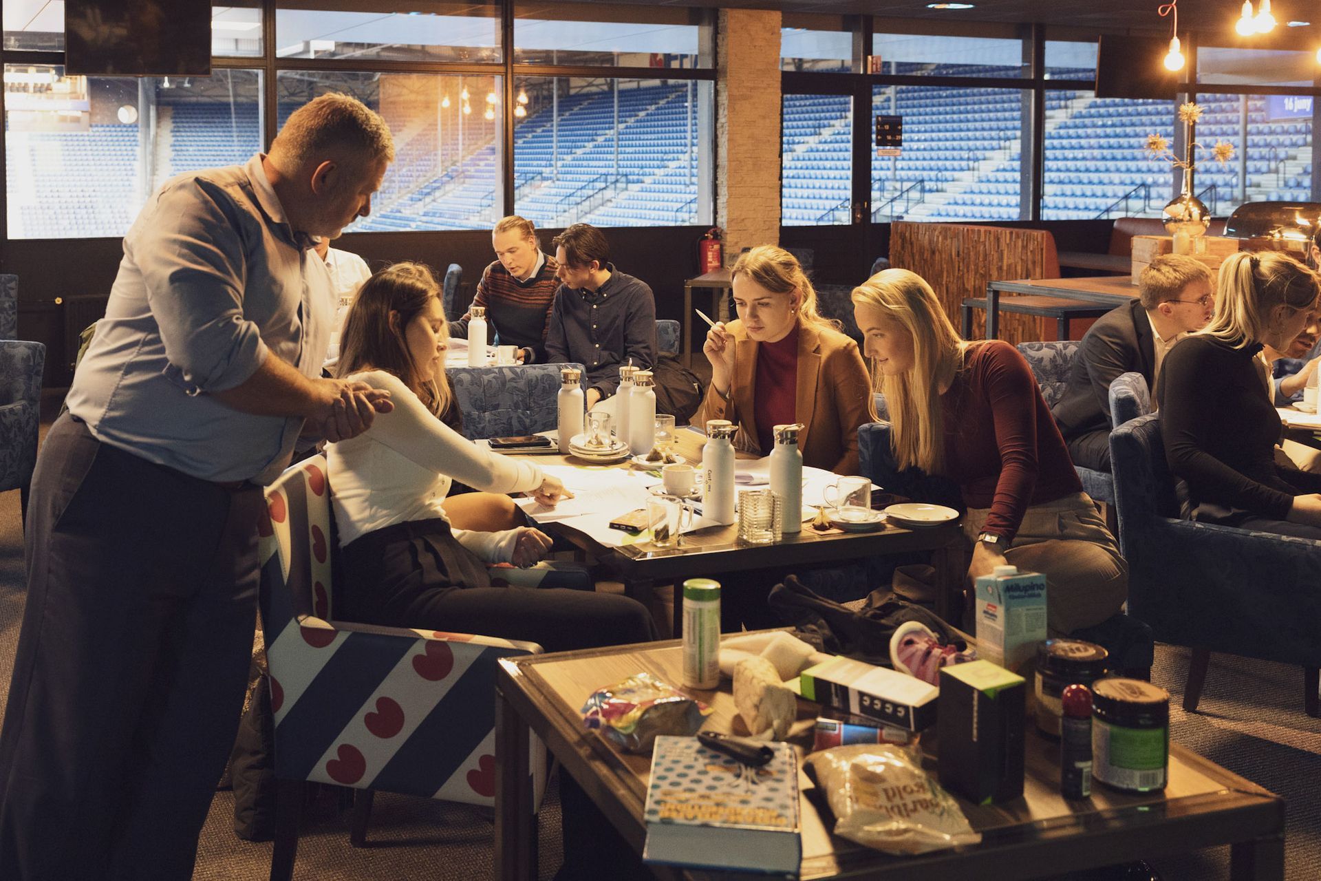Een groep mensen zit aan tafels, sommigen werken en praten. Een man helpt een vrouw die aan een tafel zit.
