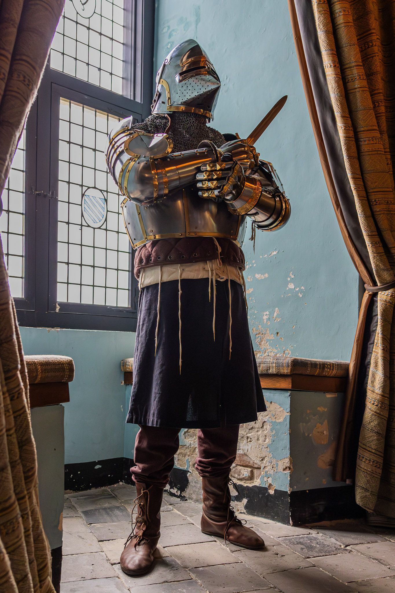 Een persoon in een volledig harnas met een zwaard in de hand. Binnen, vlakbij een raam.