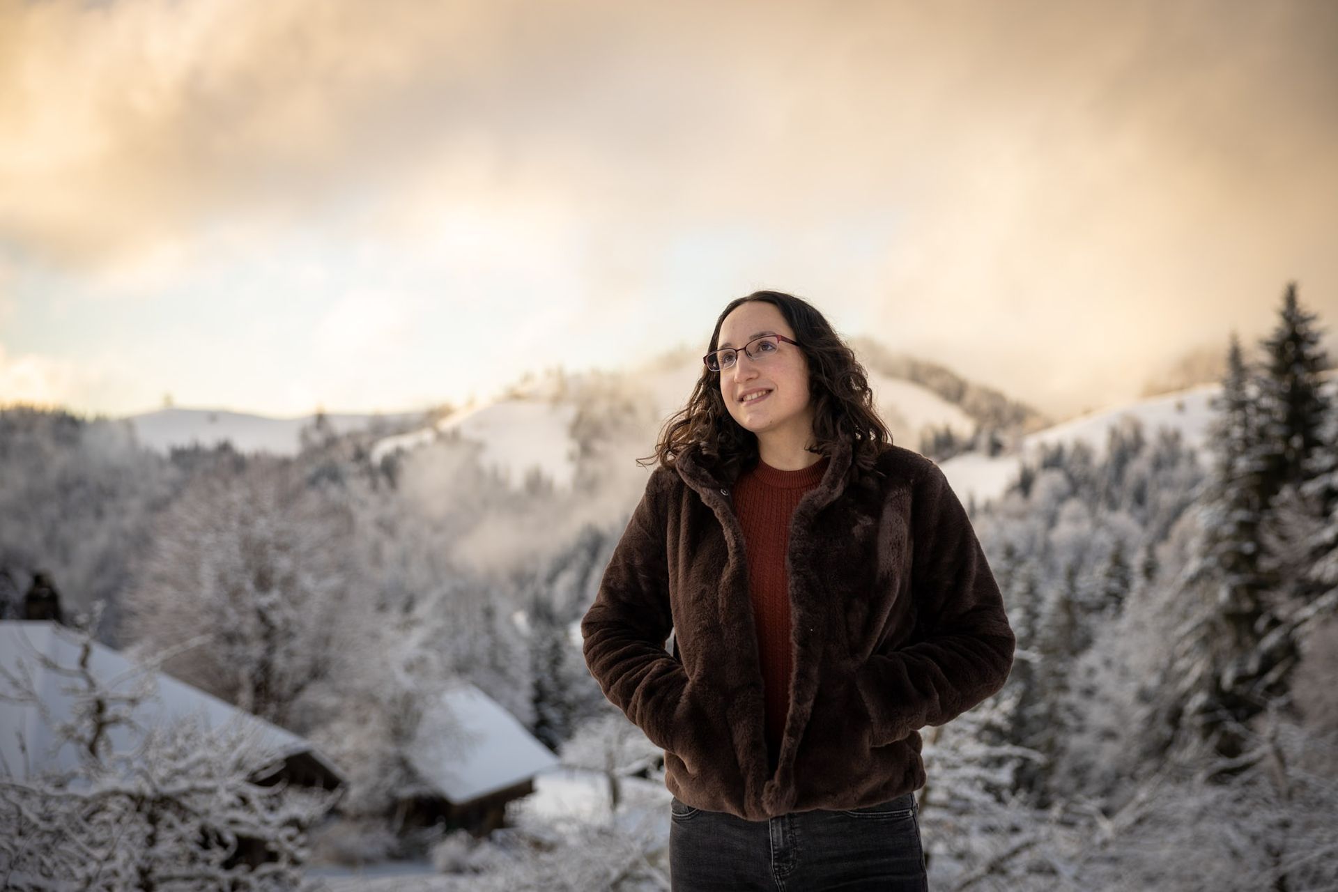 Een persoon in een bontjas staat in de met sneeuw bedekte bergen en kijkt met een glimlach weg van de camera. Bewolkte lucht.