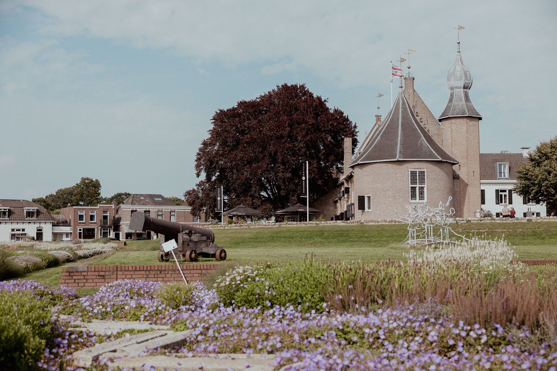 Kanon voor een stenen kasteel, omgeven door paarse bloemen en groen, op een zonnige dag.