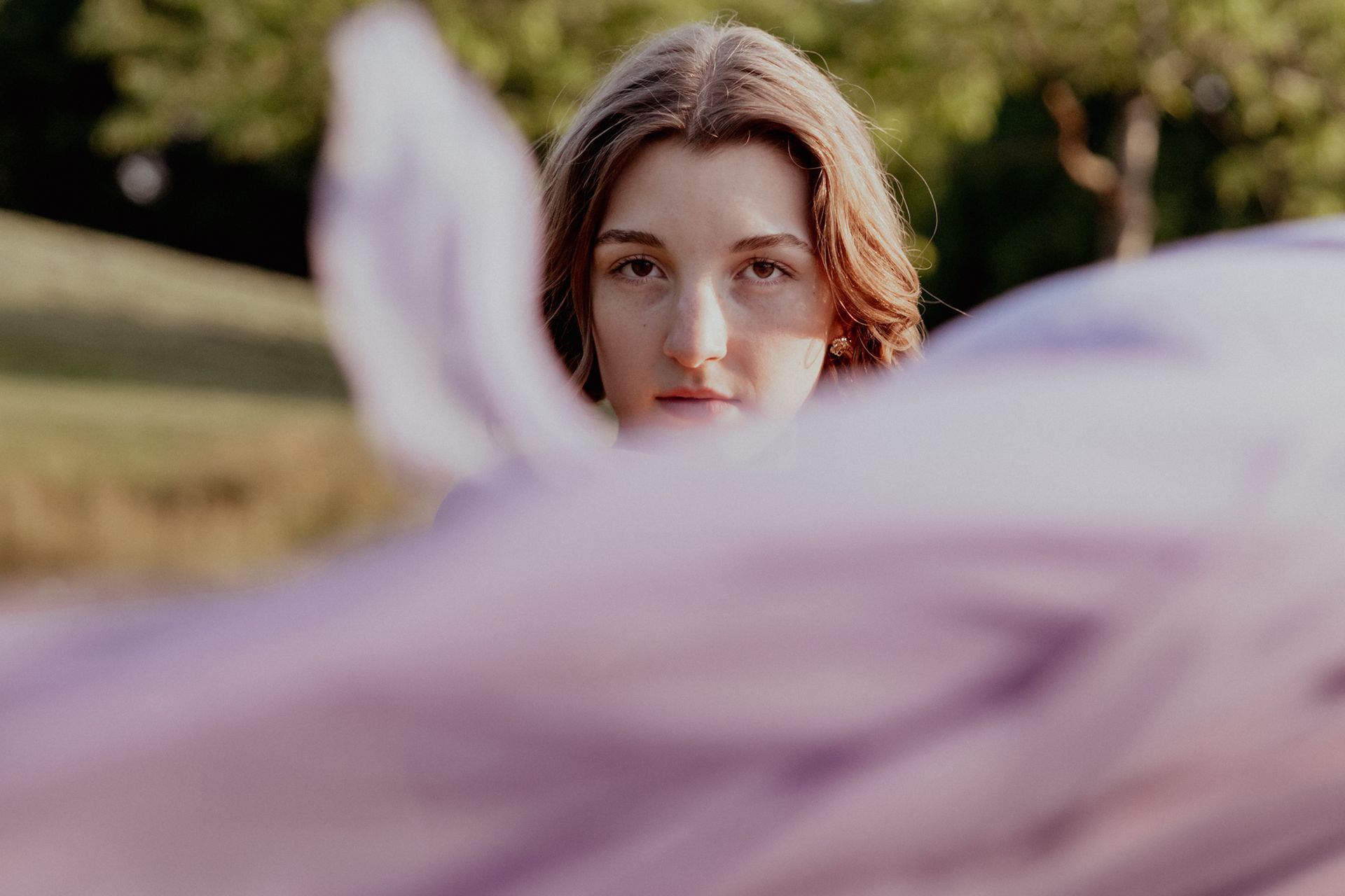 Vrouw kijkt recht in de camera, lichtpaarse stof op de voorgrond, buitenomgeving.