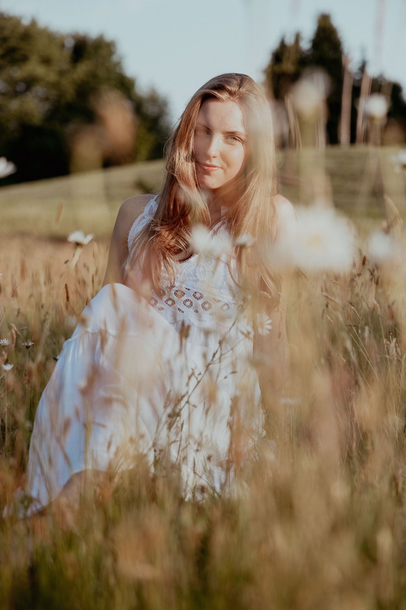 Een vrouw in een witte jurk zit in het hoge gras en lacht naar de camera. Het zonlicht baadt in het veld.
