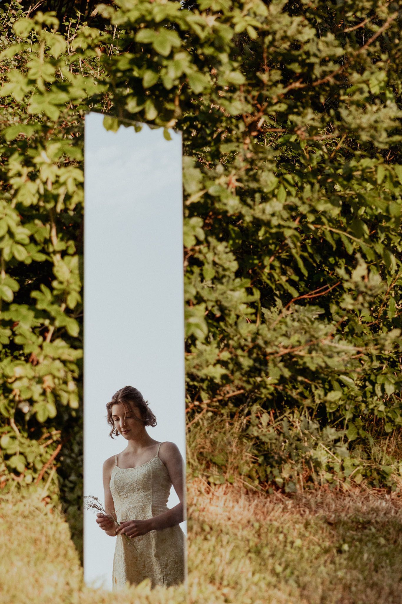 Een vrouw in een witte jurk staat voor een hoge spiegel buiten, omringd door groen gebladerte.