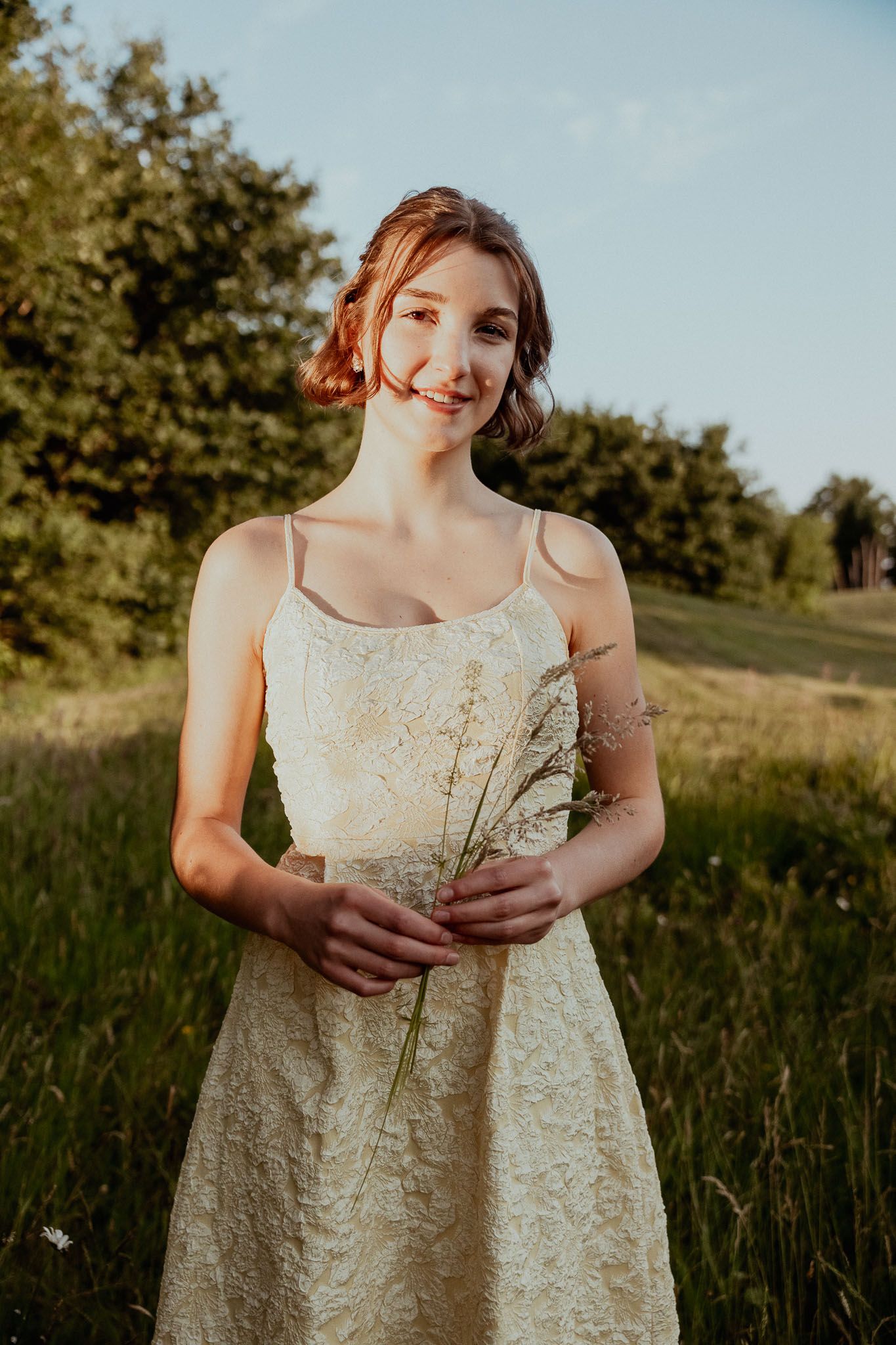 Een vrouw in een glinsterende crèmekleurige jurk, met bloemen in haar handen, lacht in een zonnig veld.
