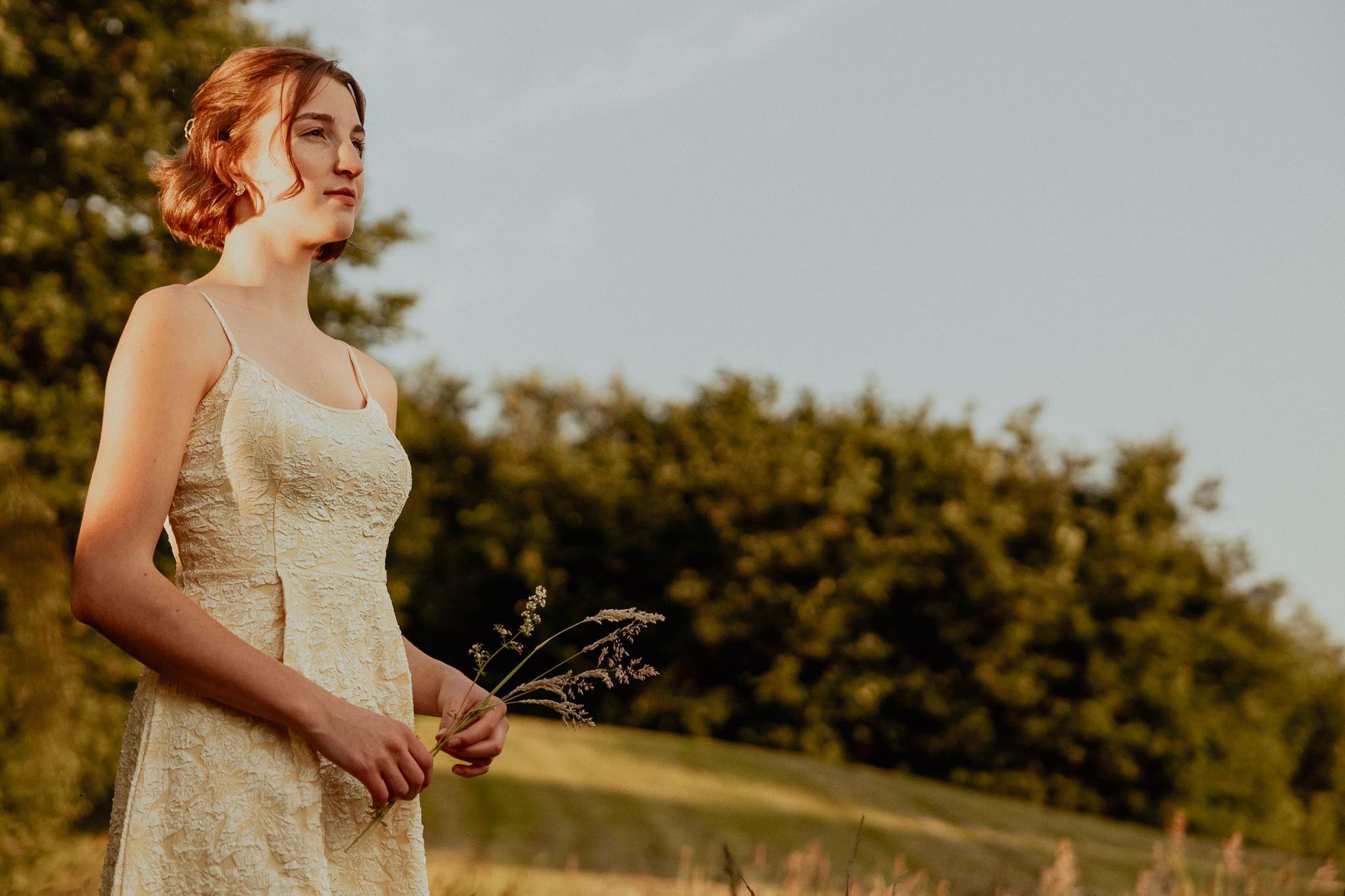 Een vrouw in een kanten jurk houdt gras vast en kijkt naar de lucht in een veld. Warm zonlicht.