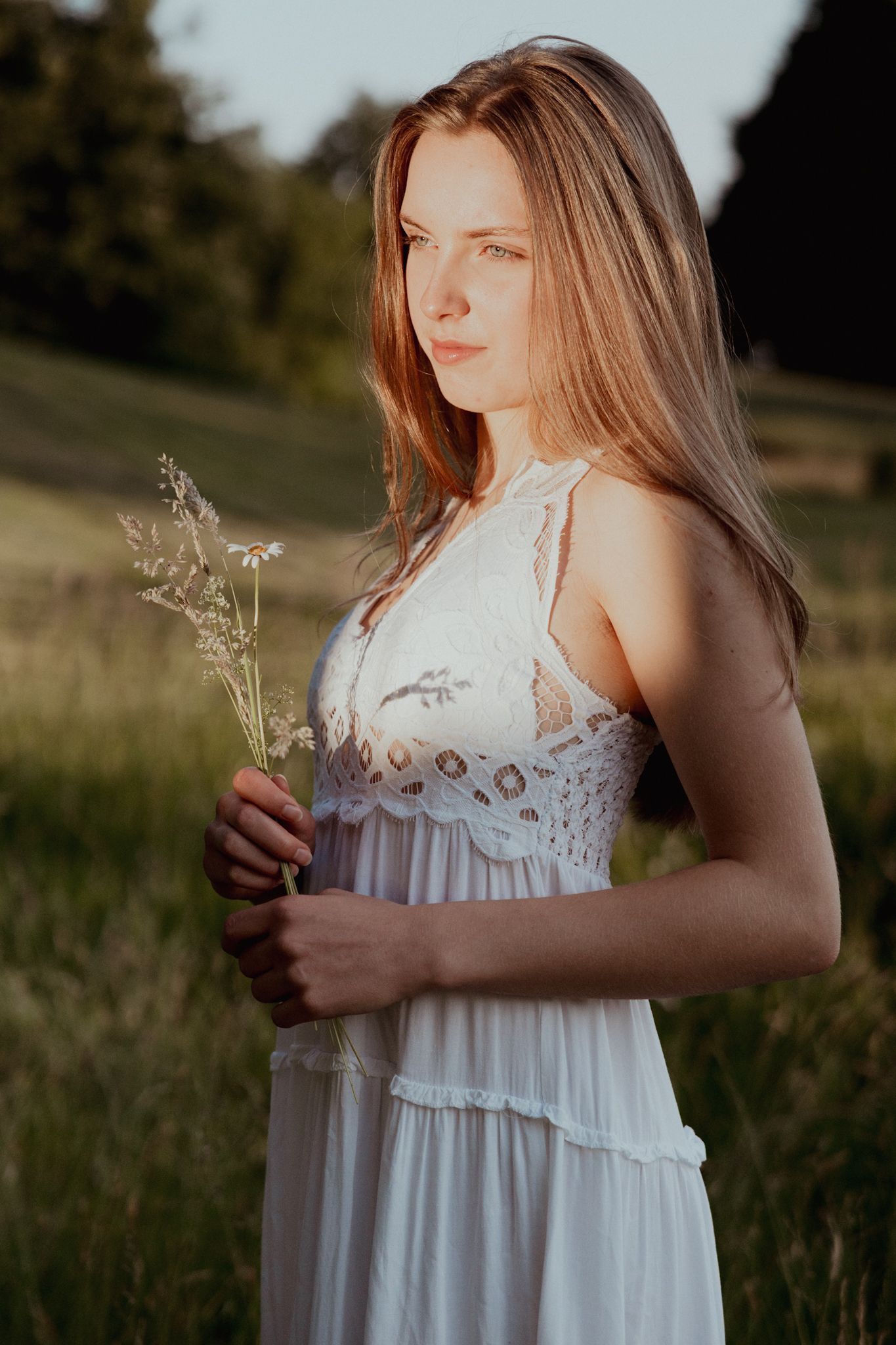 Een vrouw in een witte jurk houdt bloemen vast en staat in een zonnig veld.