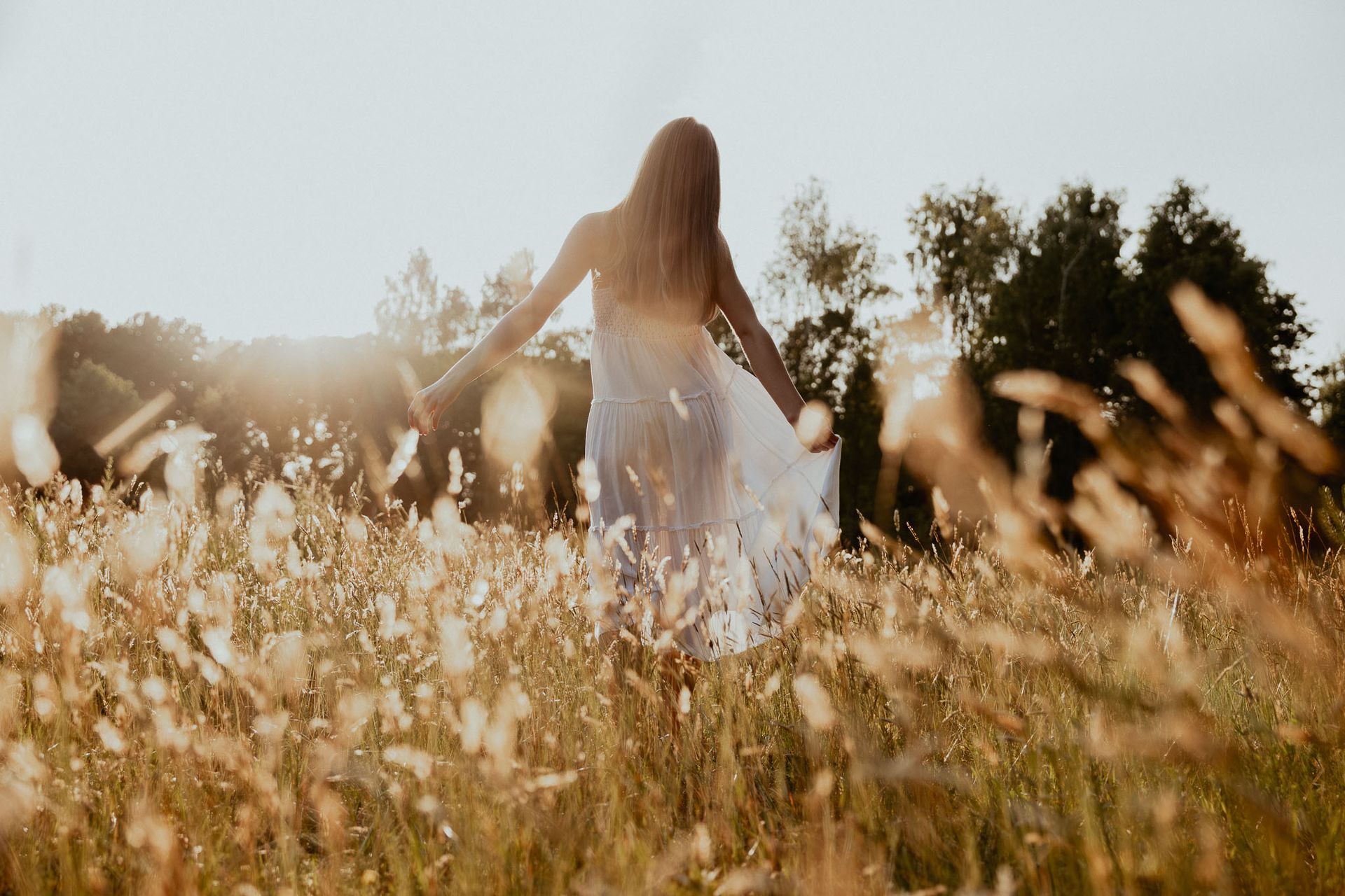 Een vrouw in een witte jurk houdt gedroogd gras vast en staat in een veld met bomen op de achtergrond. Ze kijkt in de verte.