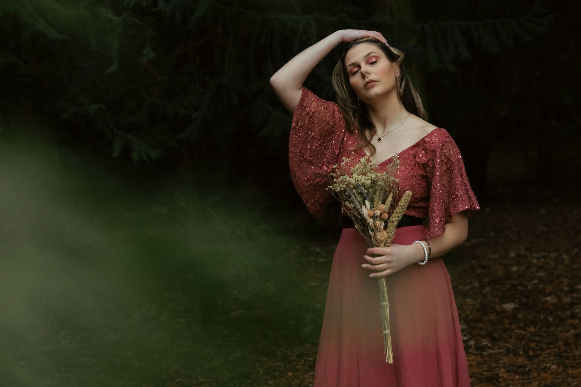 Vrouw in rode jurk houdt bloemen vast en poseert in het bos met haar hand op haar hoofd.