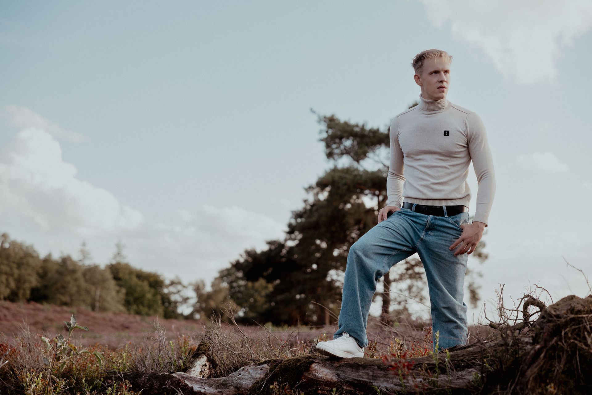 Een man in een beige shirt en spijkerbroek staat buiten en kijkt niet in beeld.