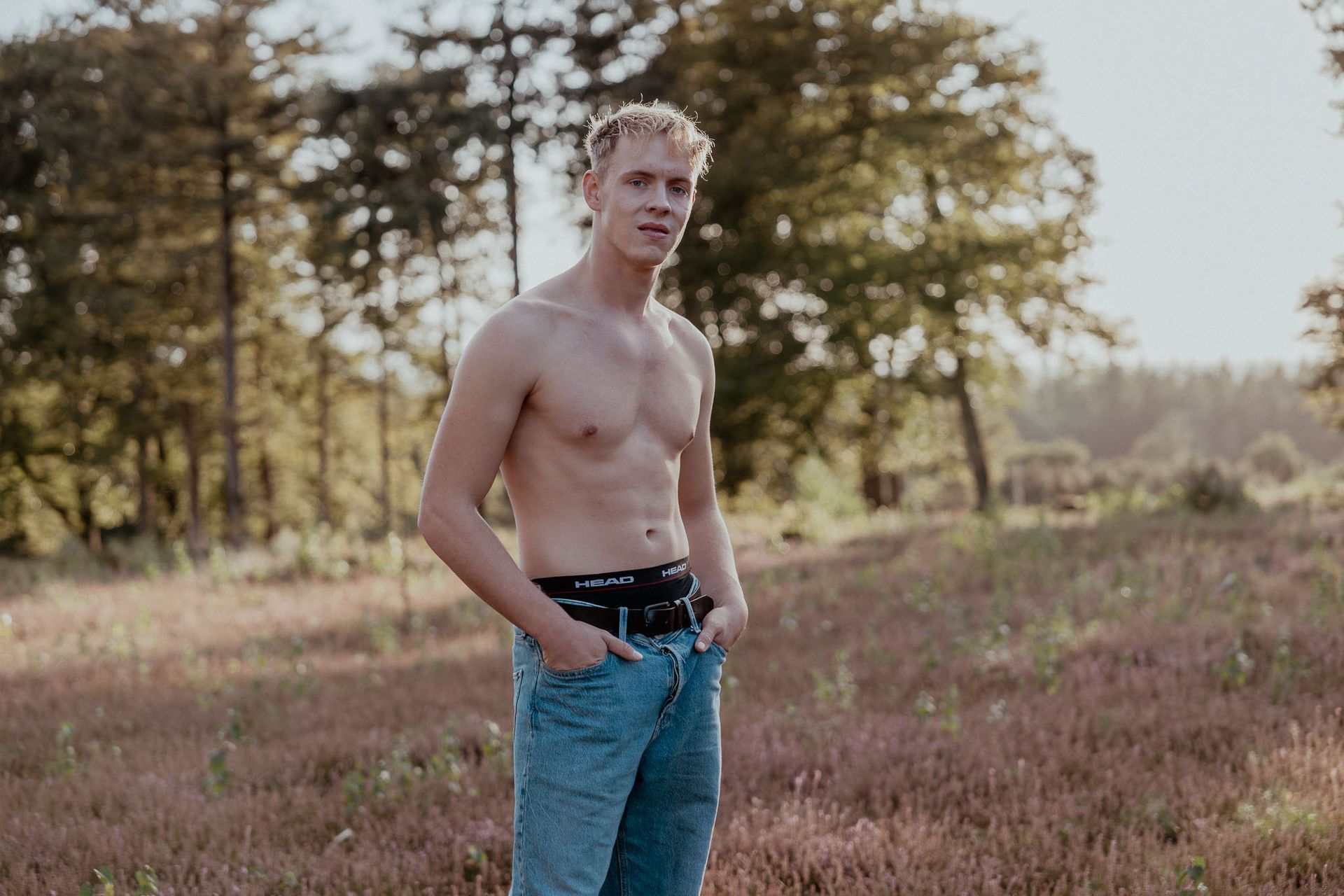 Blond persoon buiten, zonder shirt, handen in de zakken, in spijkerbroek en met een zwart heuptasje. Veld en bomen op de achtergrond.