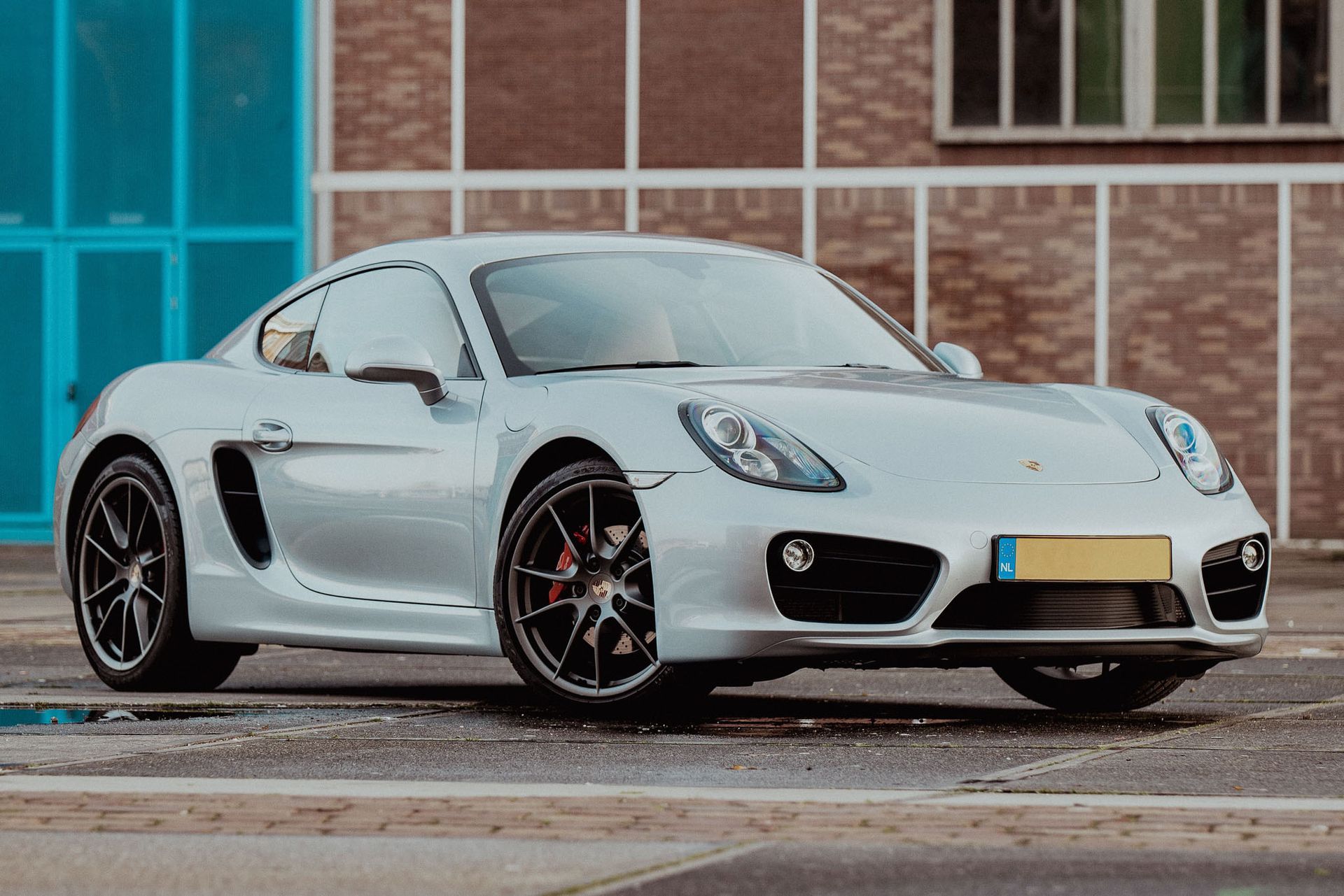 Zilveren Porsche coupé geparkeerd voor een gebouw met blauwe en bruine panelen.