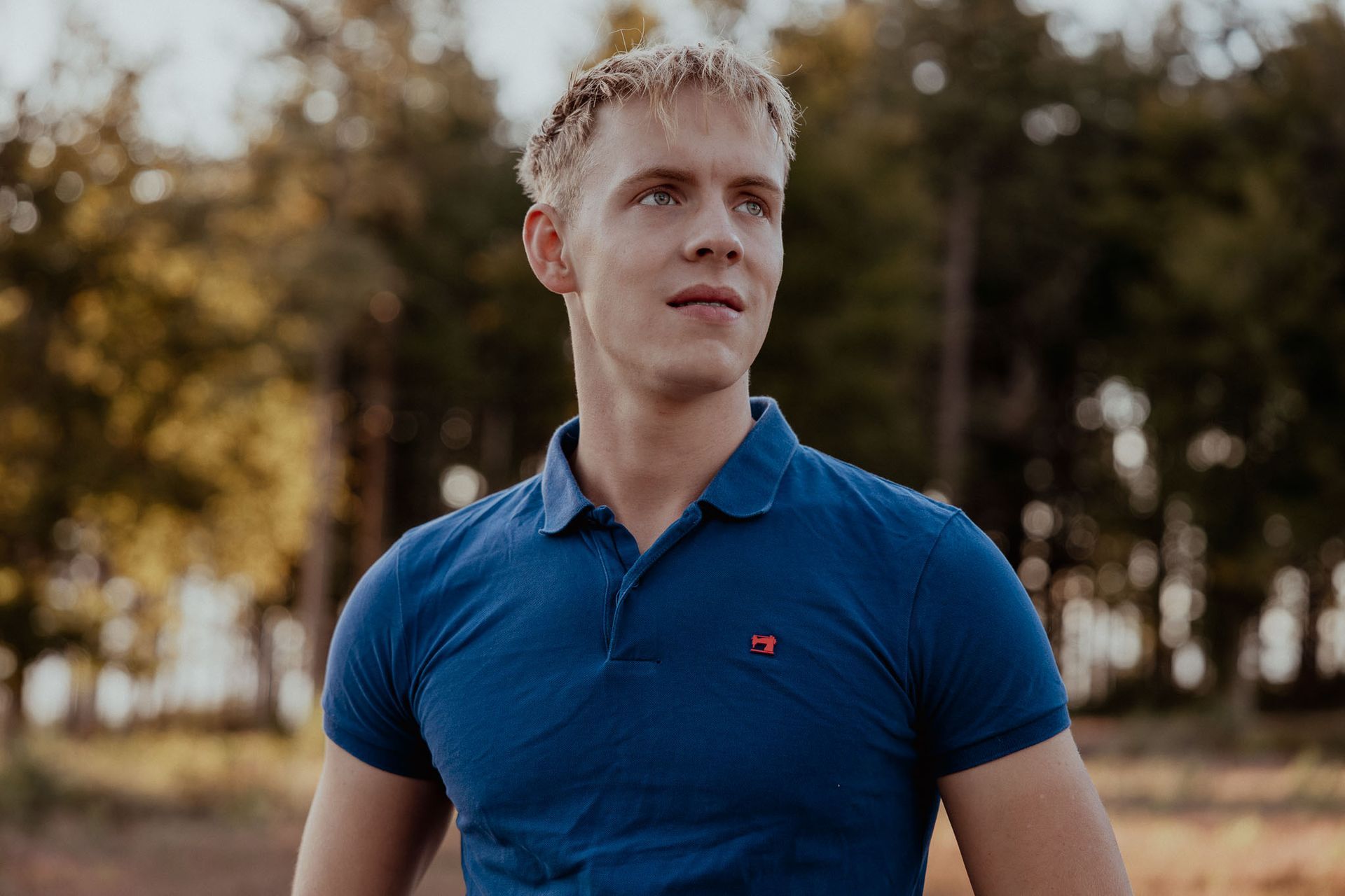 Blond persoon in een blauw poloshirt kijkt buiten beeld in een parkachtige omgeving met bomen.