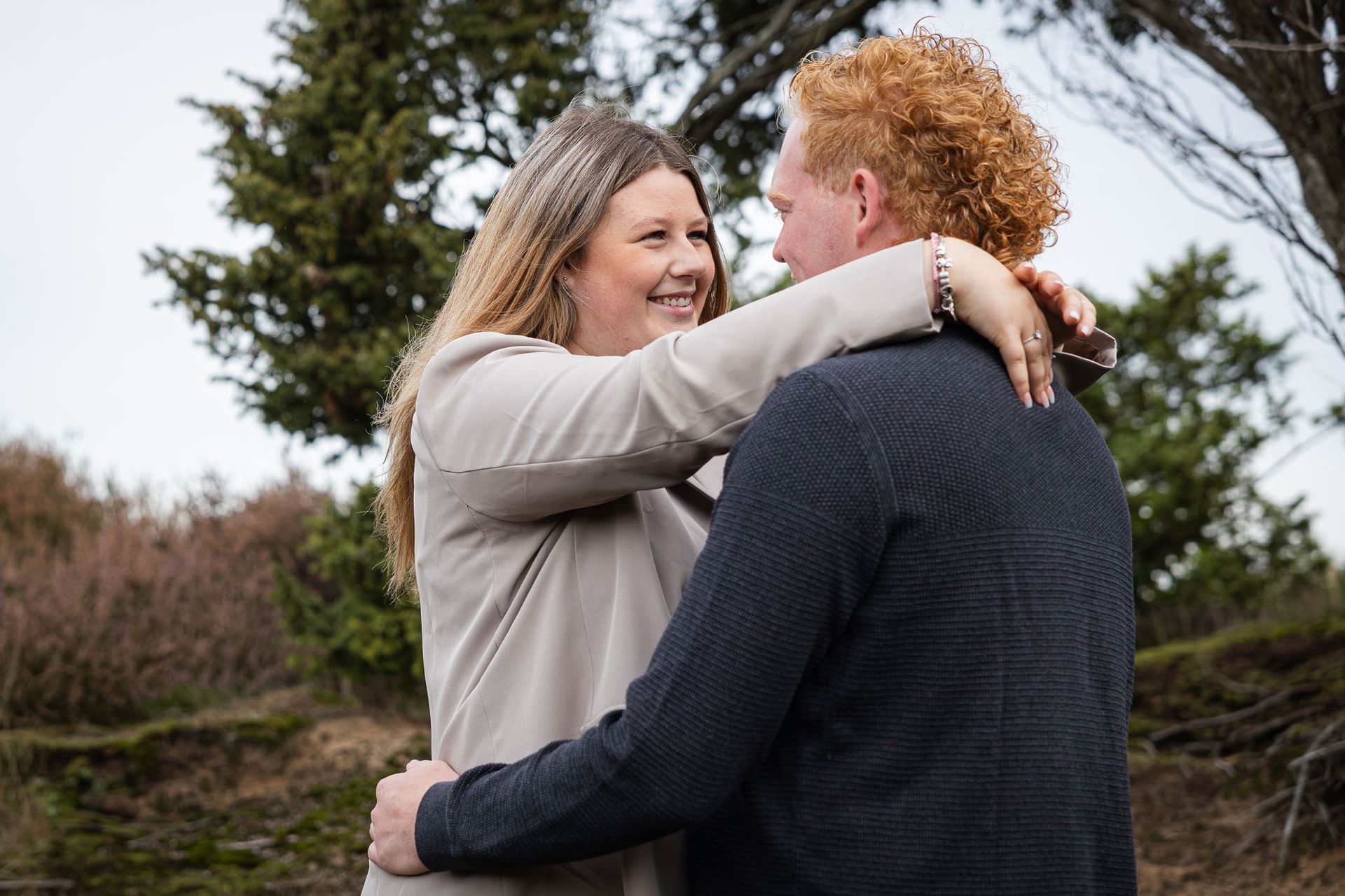 Een stel omhelst elkaar buiten, lachend. De vrouw draagt ​​een bruine jas en de man een donkere trui.