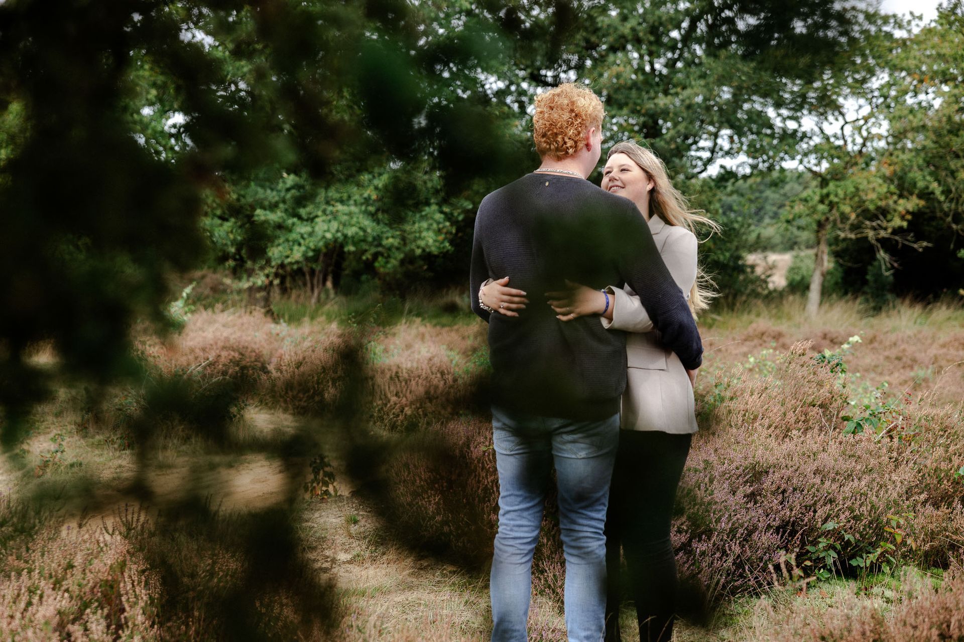 Een paar omhelst elkaar buiten, omringd door bomen en gebladerte. Man in blauwe trui, vrouw in blazer.