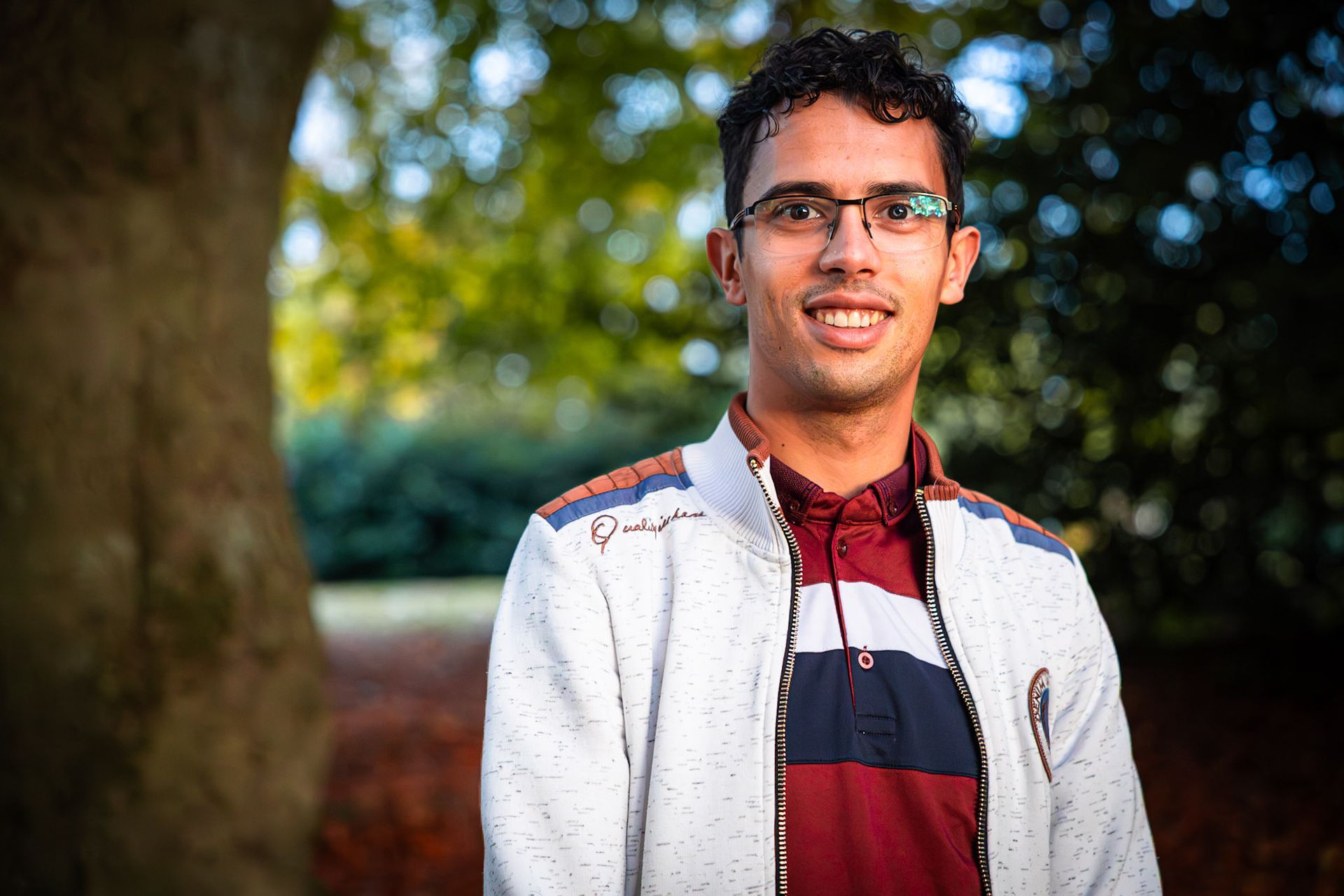 Een man met een bril en een witte jas lacht buiten, met bomen op de achtergrond.
