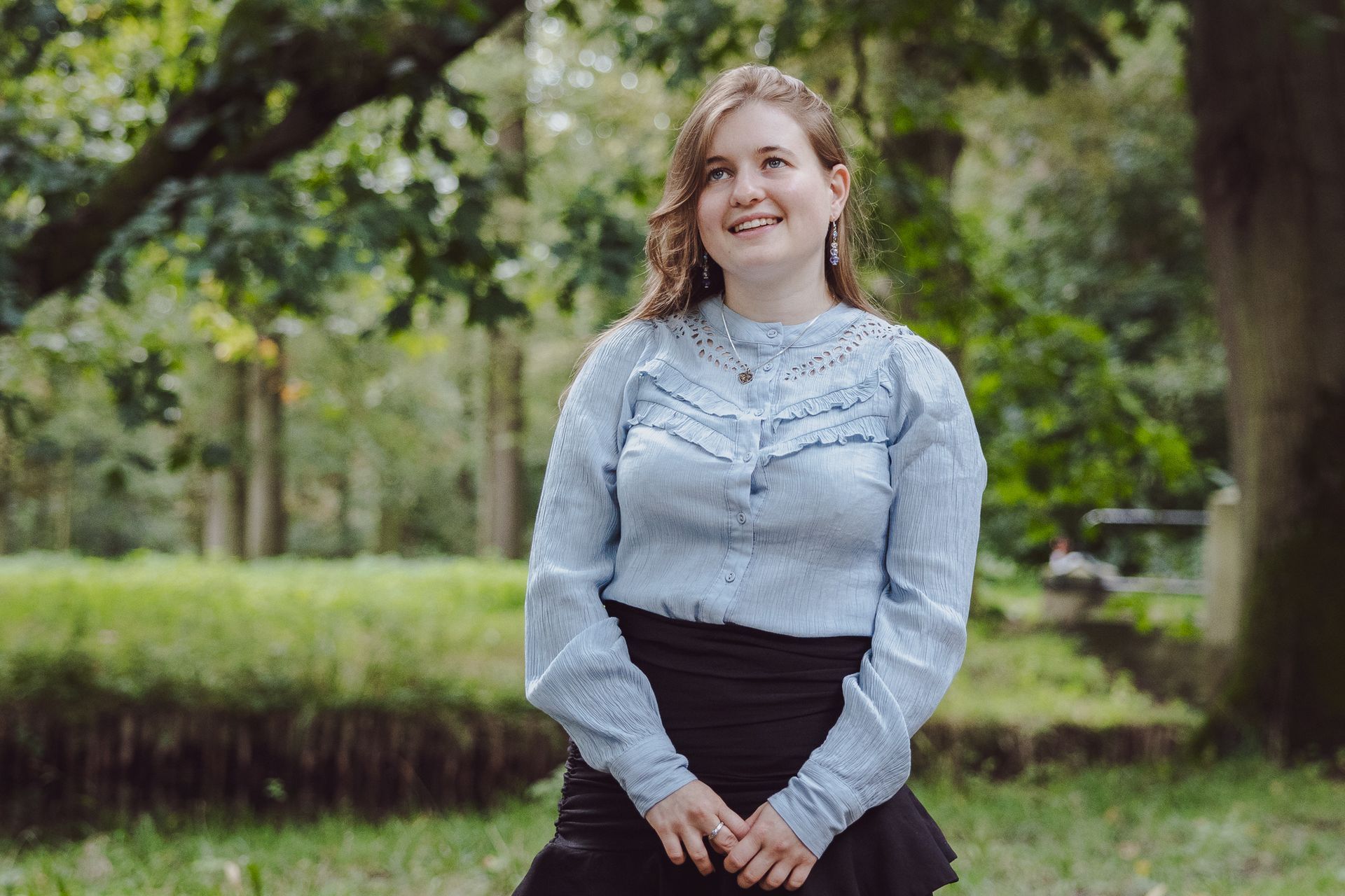 Vrouw in een blauw shirt en een zwarte rok lacht in een park.