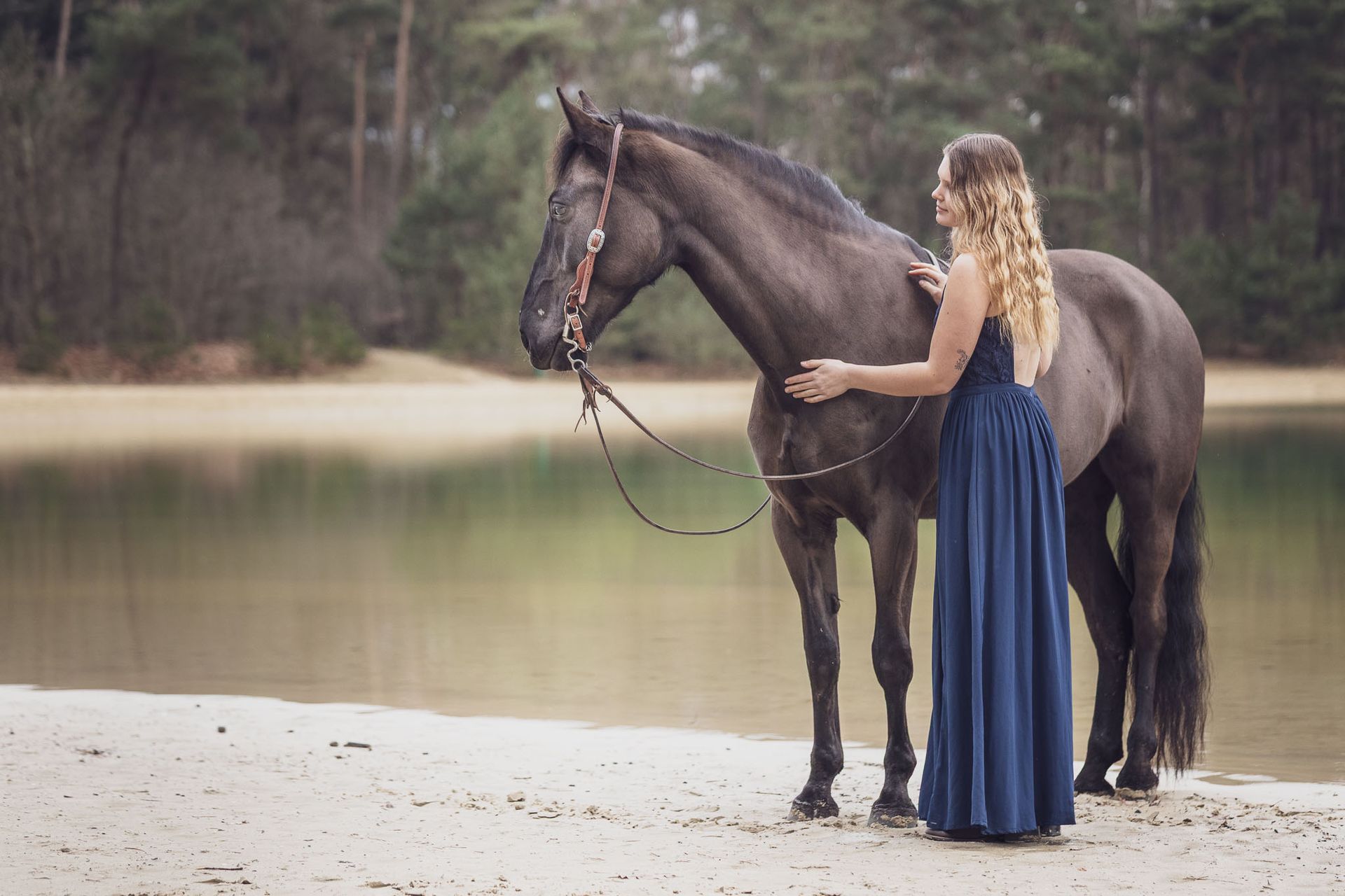 Vrouw in blauwe jurk aait een donker paard bij een meer.