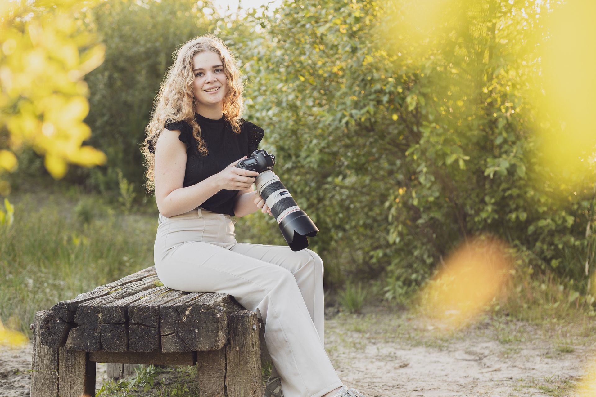 Een vrouw met krullend haar zit buiten, met een camera met een telelens in haar hand, en lacht.