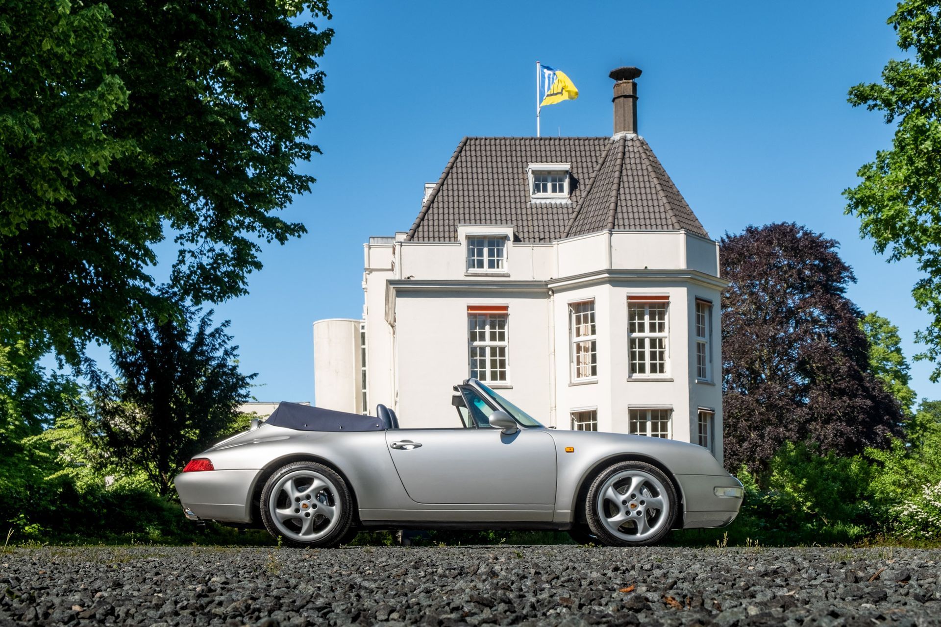 Zilveren Porsche-cabriolet geparkeerd voor een wit gebouw met bomen en een blauwe lucht.