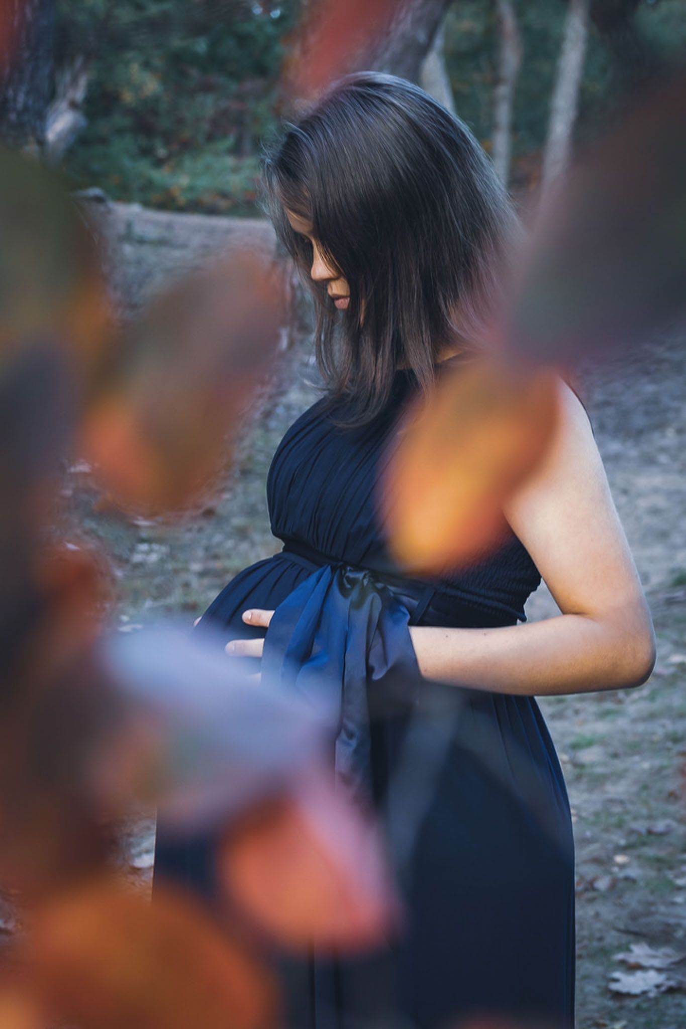 Een zwangere vrouw draagt ​​een donkerblauwe jurk en houdt haar buik vast in een natuurlijke omgeving, met bladeren op de voorgrond.