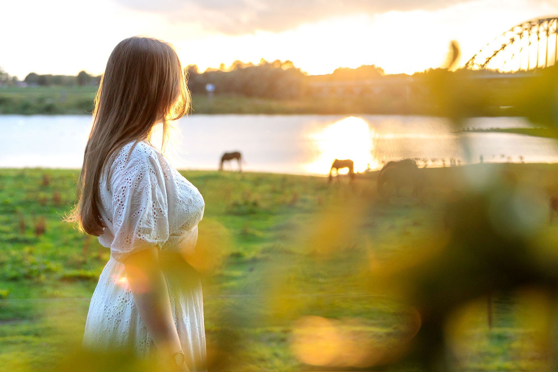 Een vrouw in een witte jurk kijkt naar de zonsondergang boven het water, met in de verte twee paarden.