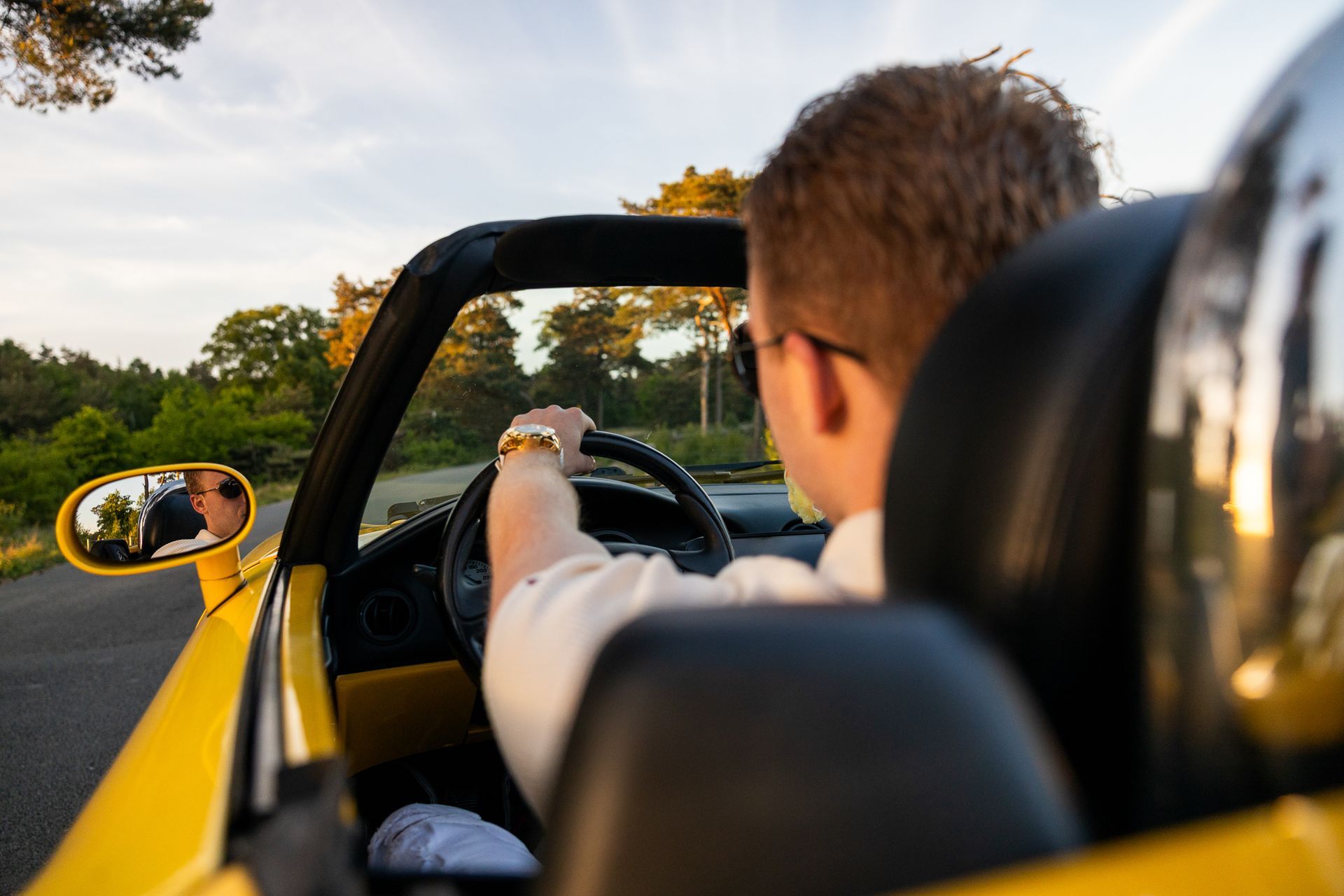 Man rijdt in een gele cabriolet over een met bomen omzoomde weg bij zonsondergang. Hij draagt ​​een zonnebril en een gouden horloge.