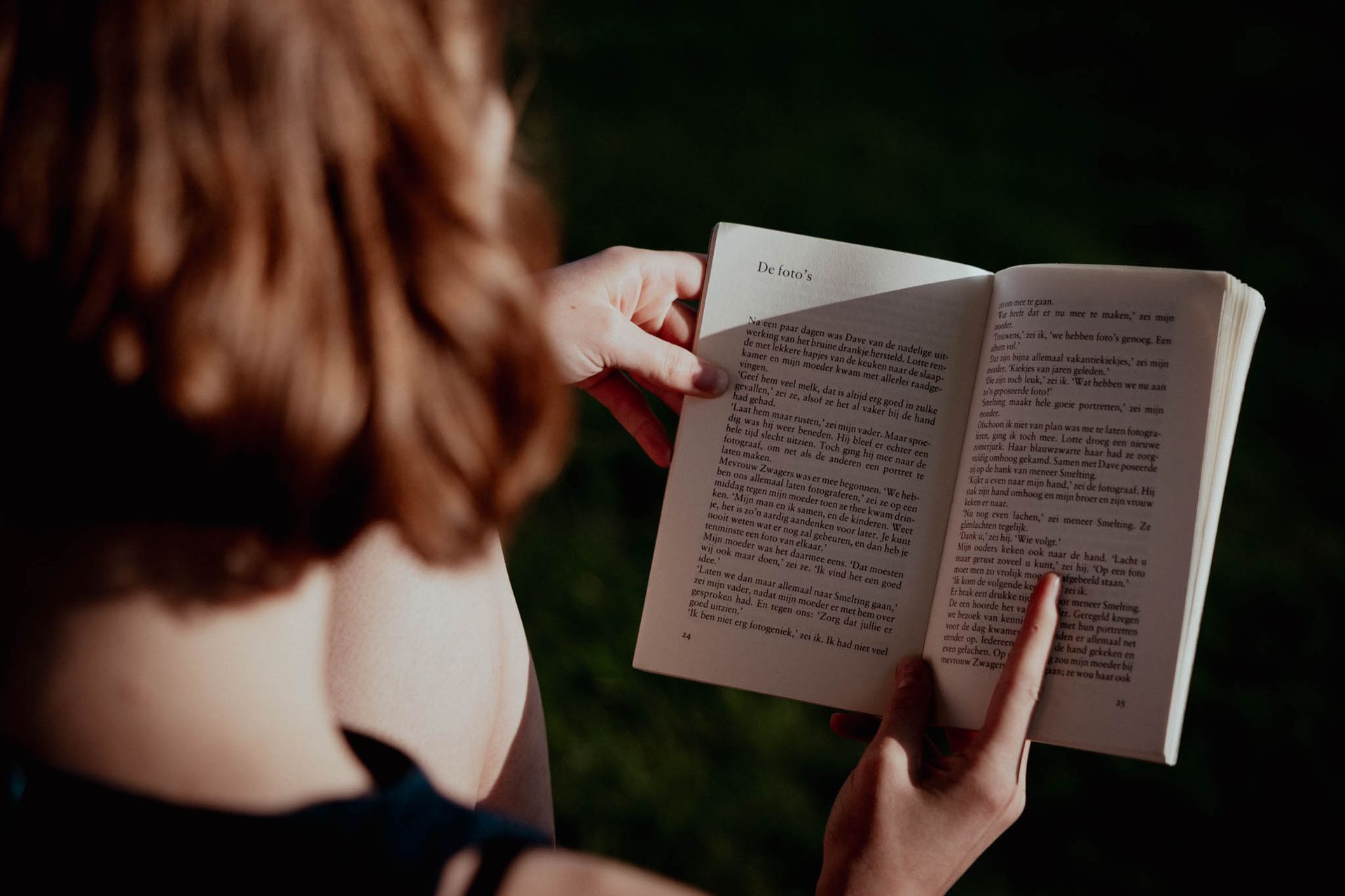 Een persoon met rood haar leest een boek in de buitenlucht, in de zon.
