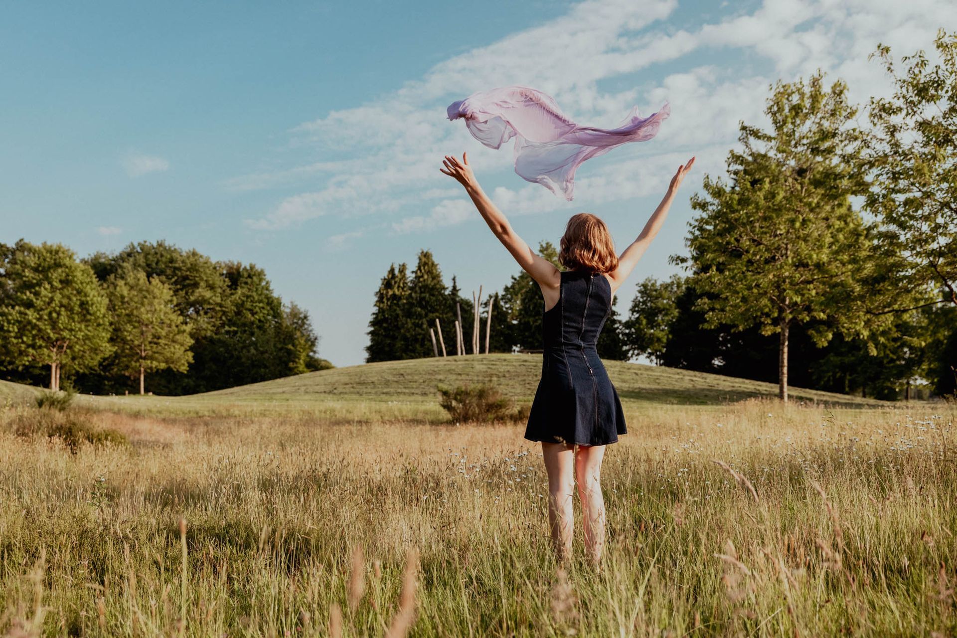Vrouw in een donkere jurk met opgeheven armen, die een sjaal in de lucht gooit, in een veld onder een blauwe hemel.