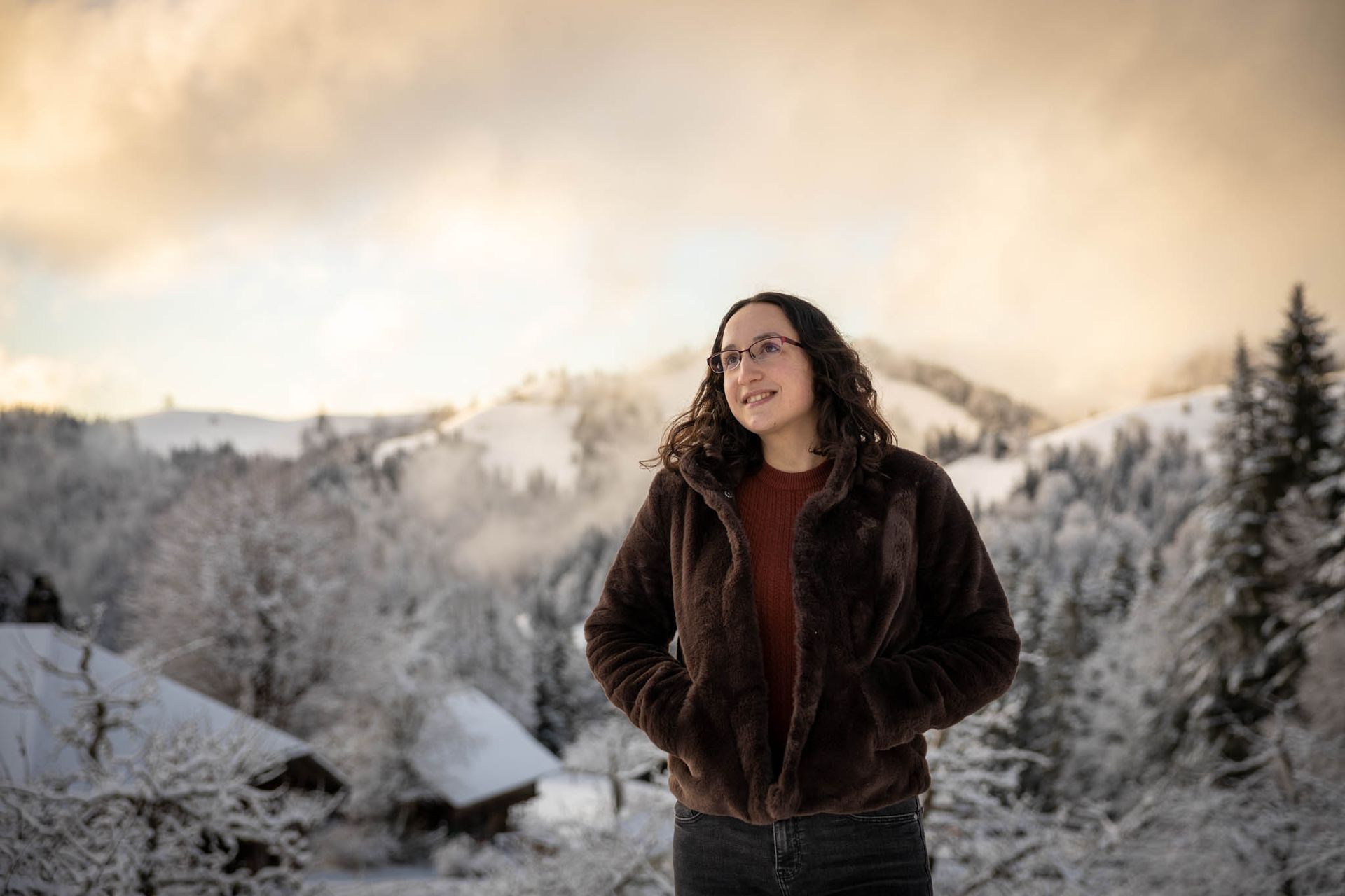 Vrouw in een bruine jas, besneeuwde bergen op de achtergrond, handen in haar zakken.