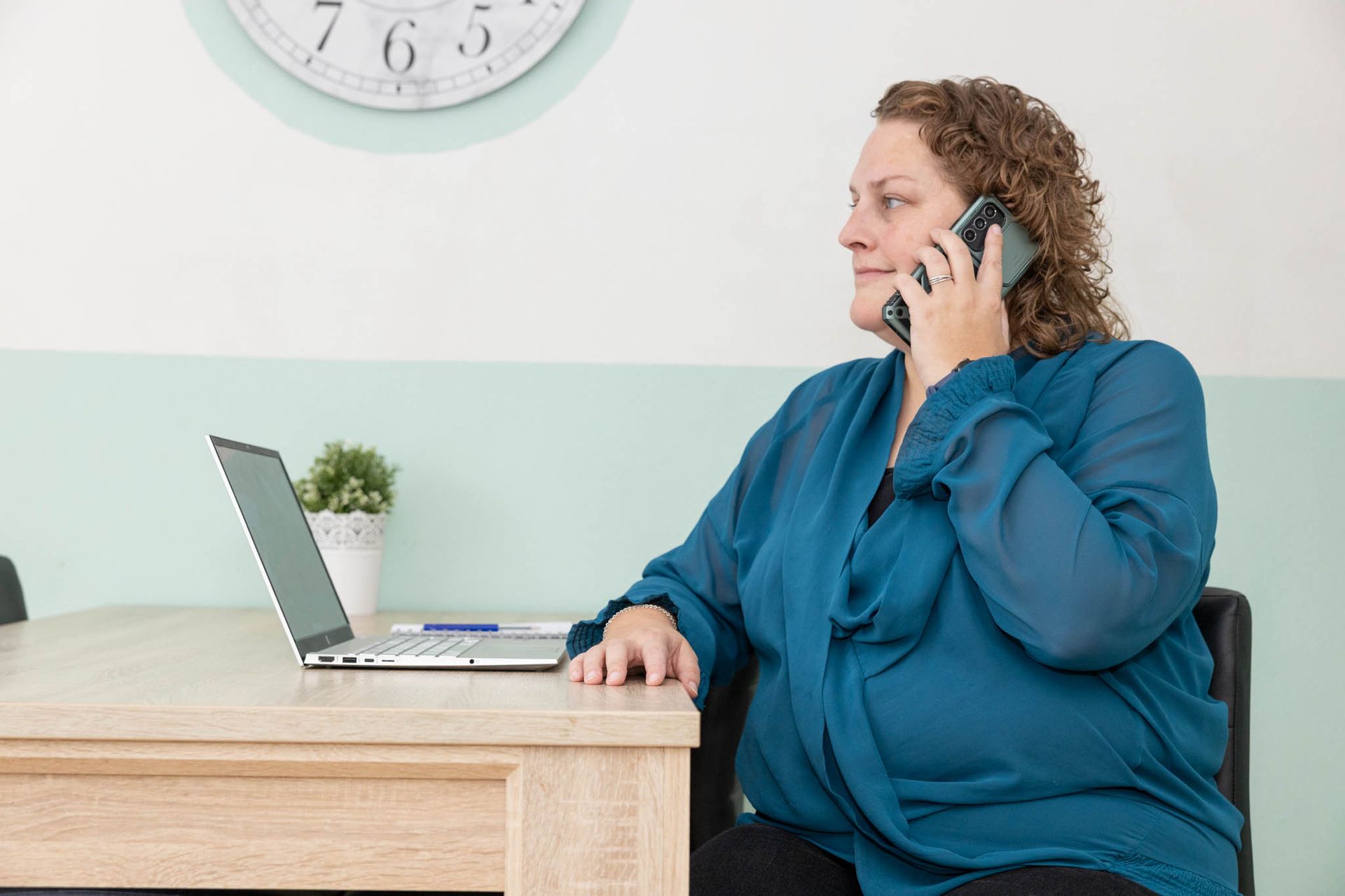 Vrouw aan de telefoon, zittend aan een bureau met een laptop. Achter haar hangt een klok aan de muur.
