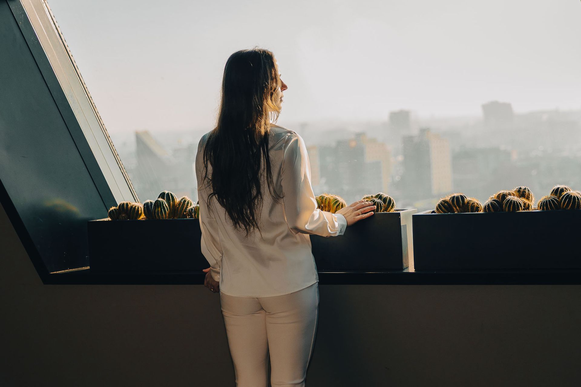 Een vrouw kijkt door een groot raam naar een stadsgezicht, verlicht door de zon.