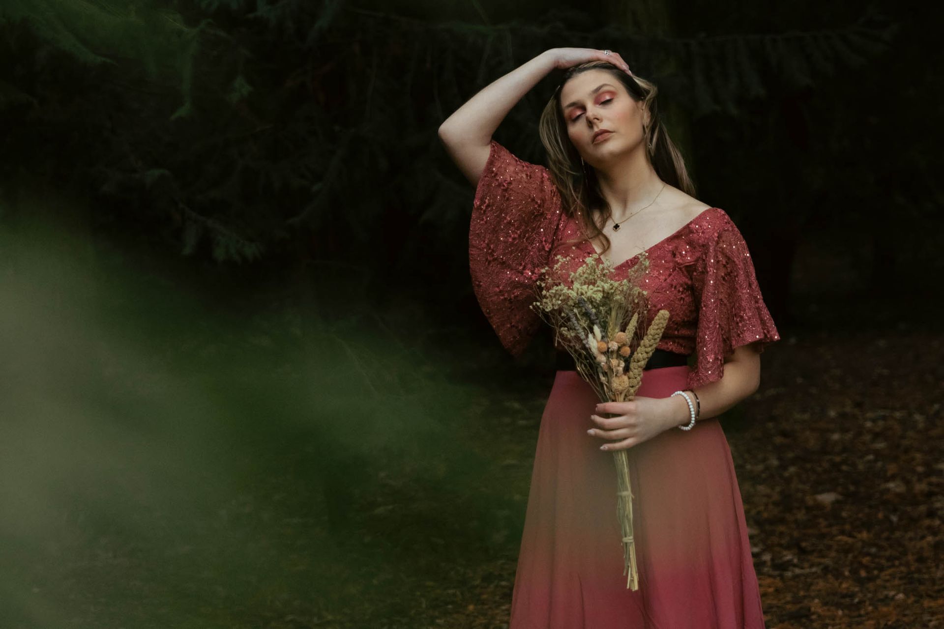 Een vrouw in een rode jurk met mouwen van pailletten houdt een boeket vast, met haar hand op haar hoofd, in een bos.