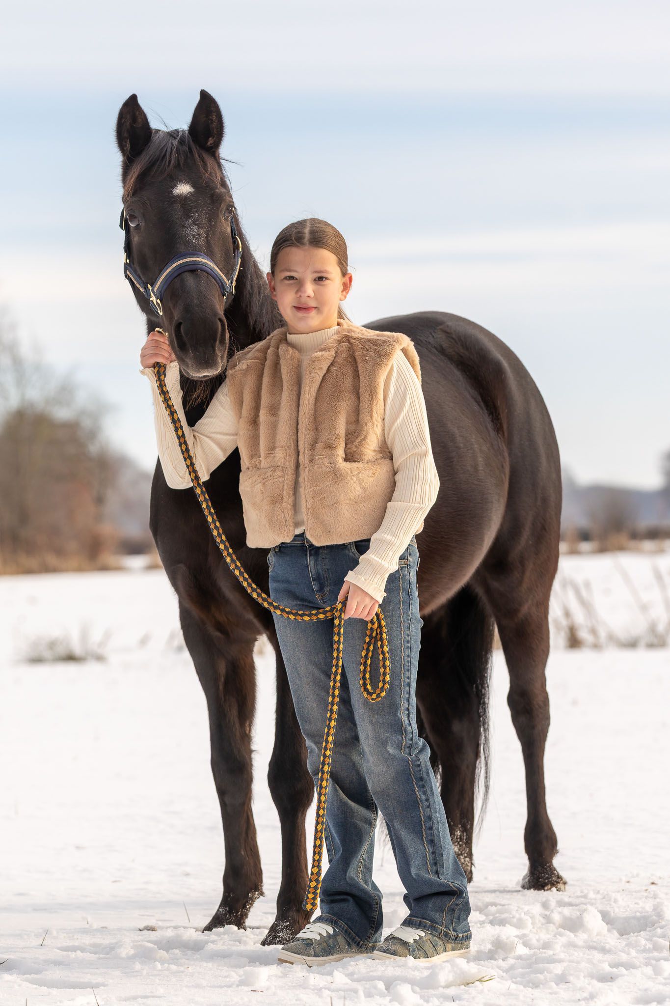 Een jong persoon in spijkerbroek en vest houdt een zwart paard vast in een besneeuwd veld.