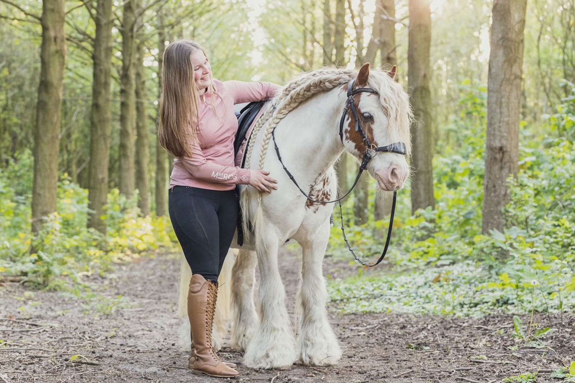 Een vrouw aait een wit paard in een bosrijk gebied; beiden hebben gevlochten manen en dragen rijtuig.