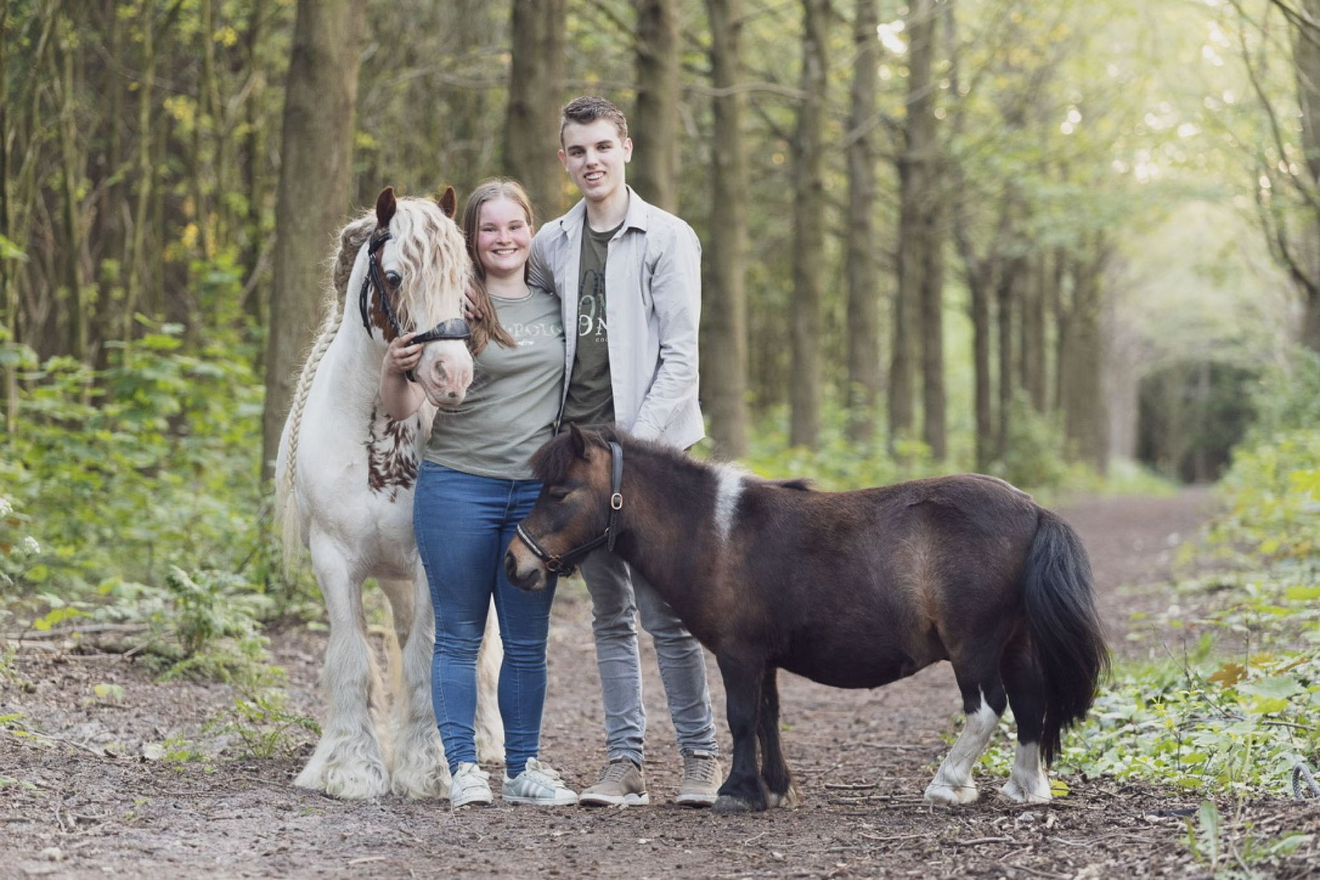 Een stel poseert met twee paarden op een bospad. Een groot wit paard en een kleine bruine pony zijn bij hen.
