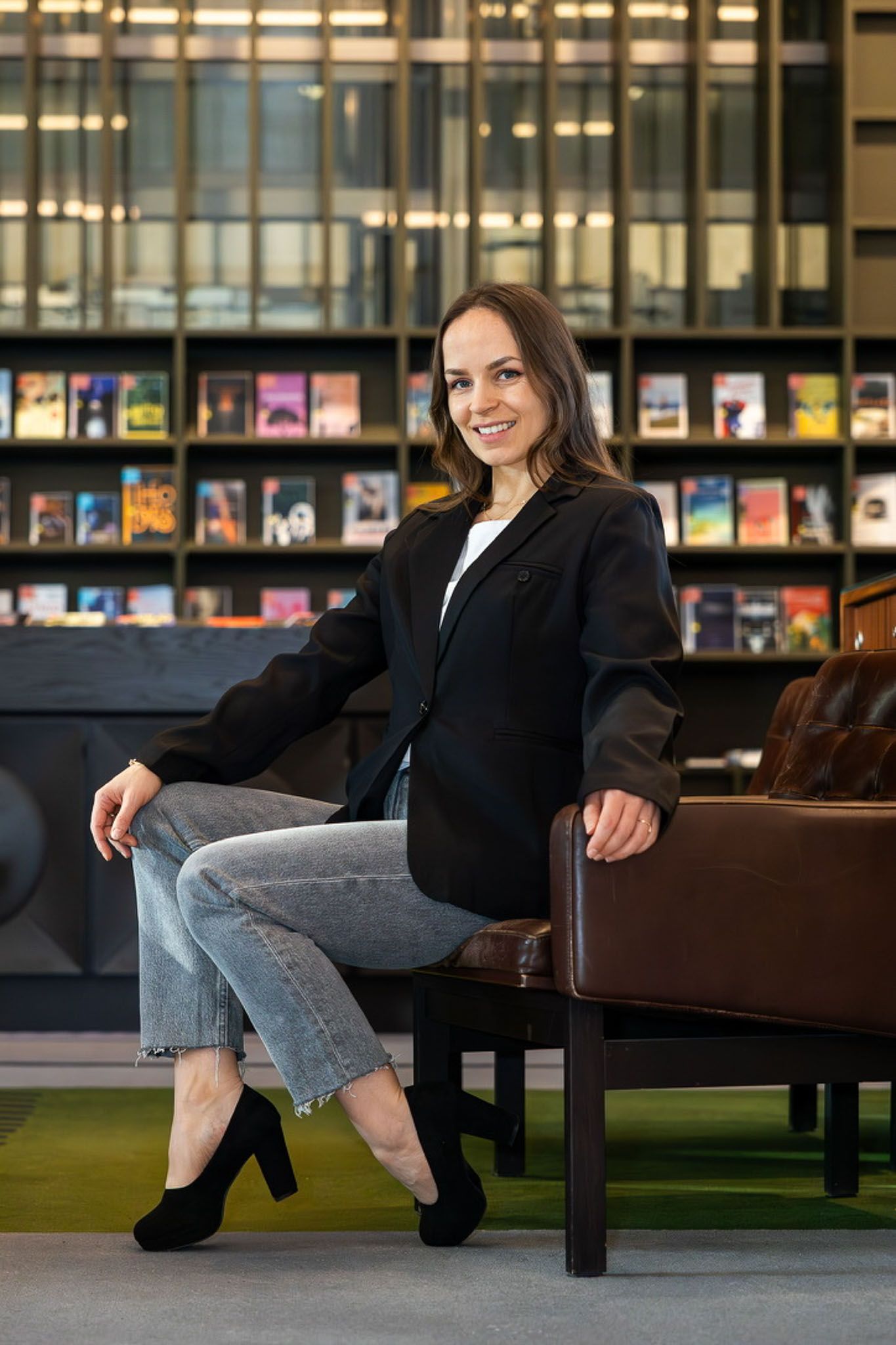 Een vrouw in een blazer en jeans zit glimlachend voor een boekenplank.